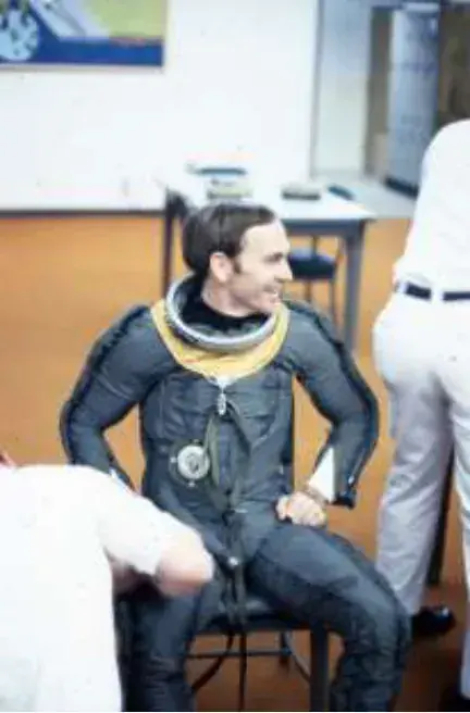A pilot in a pressure suit and helmet seated indoors
