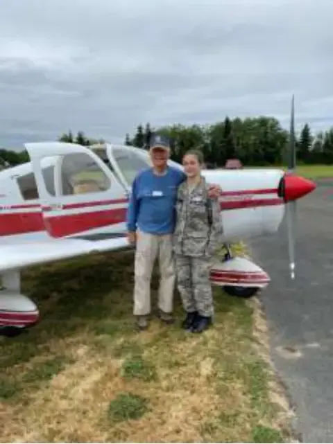 Two people standing beside a red and white airplane on grass