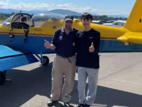 Two men giving a thumbs up in front of a blue and yellow airplane