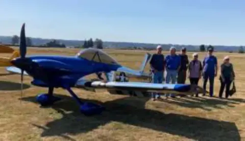 People standing by a blue homebuilt airplane on a grass field