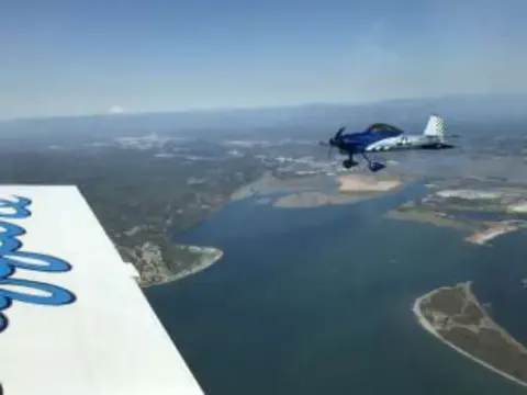 A blue and white airplane flying over water and a sandy spit, seen past a wing