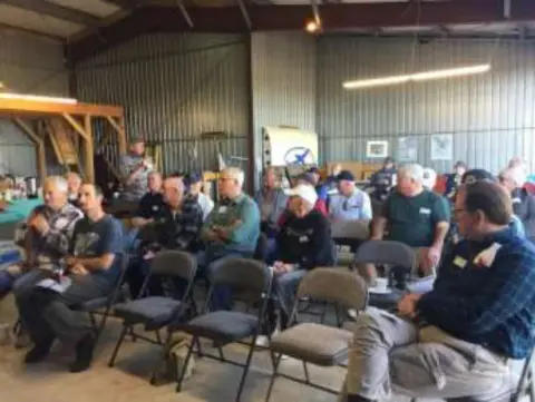 Members seated in rows of chairs in a hangar for the gathering