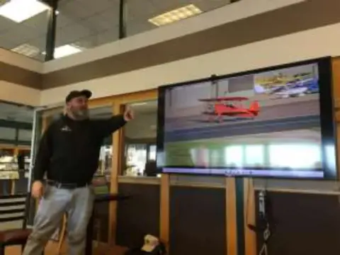 A man pointing to a screen showing a red biplane