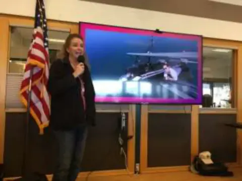 A woman speaking with a microphone beside a screen and a flag