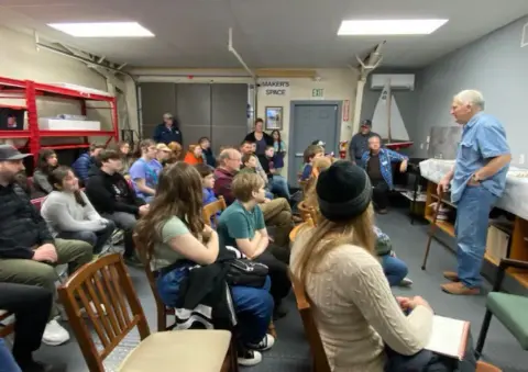 A man presenting to young people seated in a hangar classroom