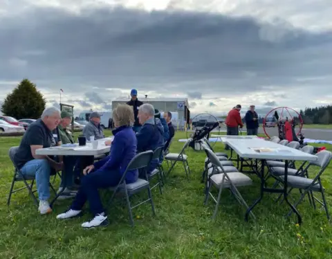 Members seated at tables on the grass beside the airfield