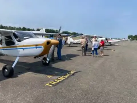 A white and yellow high-wing airplane on the ramp with people beyond