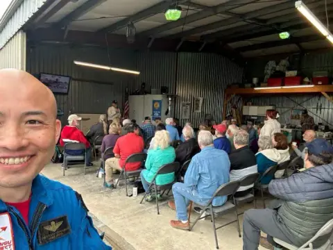 A man in a blue flight suit taking a selfie in front of an audience in a hangar