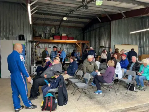 A man in a blue flight suit speaking to an audience in a hangar