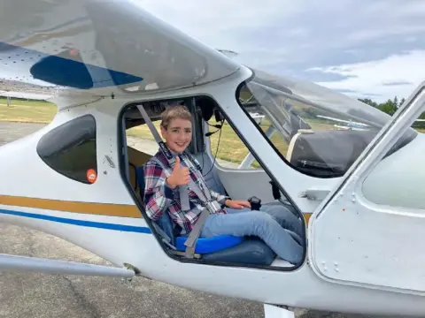A smiling woman seated in the cockpit of a white high-wing airplane