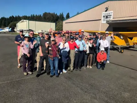 A large group of members in front of a hangar with yellow airplanes