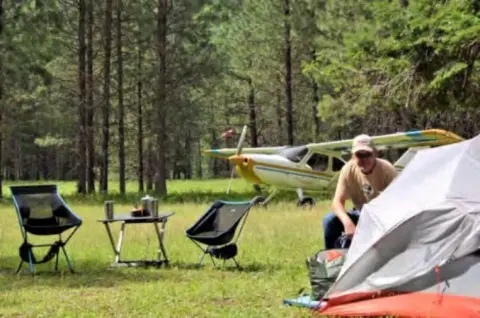 A man with camping chairs and a covered airplane in a forest clearing