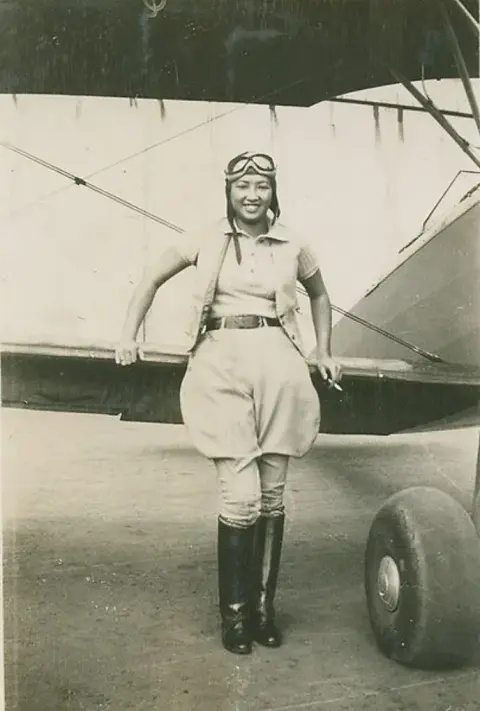 Vintage photo of a woman pilot in a flight suit standing in front of a biplane