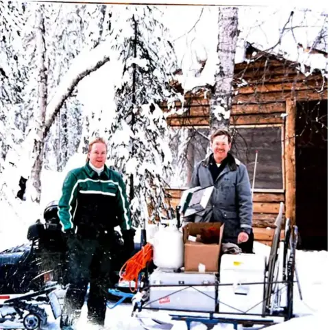 Two men in winter jackets beside a loaded sled in front of a log cabin