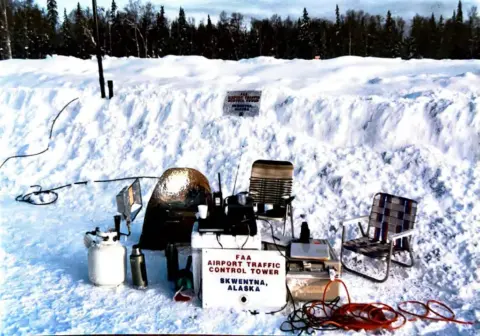 Radios, a generator, lawn chairs and a control tower sign set up beside a snowbank