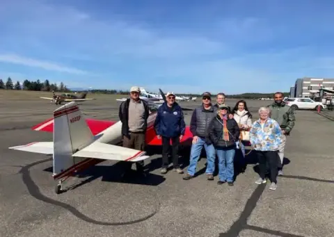 Group photo beside a blue and white airplane on the ramp, shown next to the same airplane parked