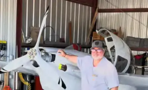 Builder leaning on the canard of a white composite airplane in a hangar