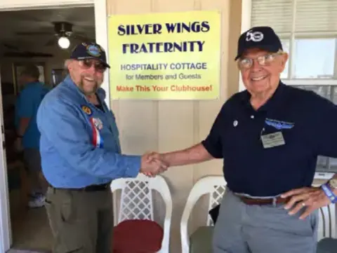 Two men shaking hands in front of a Silver Wings Fraternity hospitality cottage sign