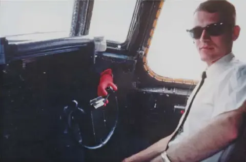 Pilot in sunglasses and tie seated in an airliner cockpit, with a red glove on the control column