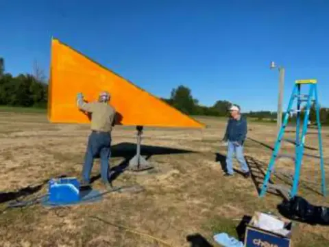 Two men painting a large orange tetrahedron wind indicator on a stepladder