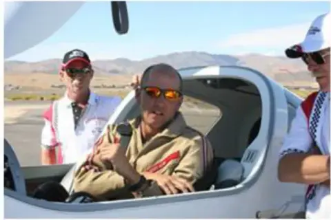 Pilot in sunglasses seated in the open cockpit of a race plane with crew beside him