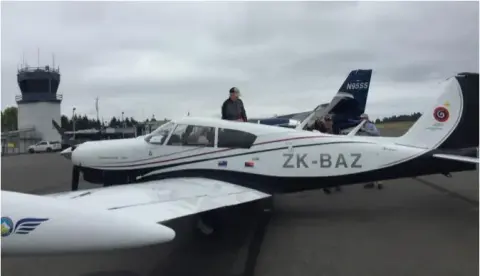 Low-wing airplane registered ZK-BAZ parked in front of a control tower