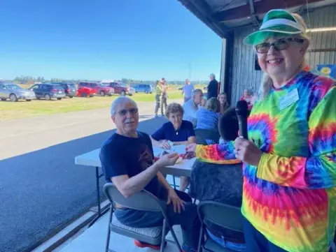 Poker game players seated at tables in an open hangar, with the host in a tie-dye shirt