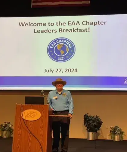 Speaker at a podium below a screen reading Welcome to the EAA Chapter Leaders Breakfast