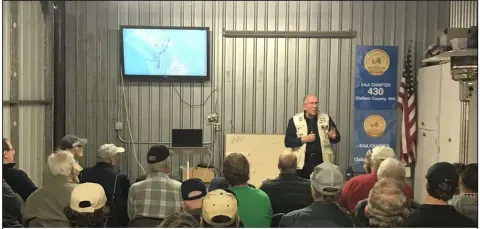 Audience in a hangar watching a presenter beside a screen