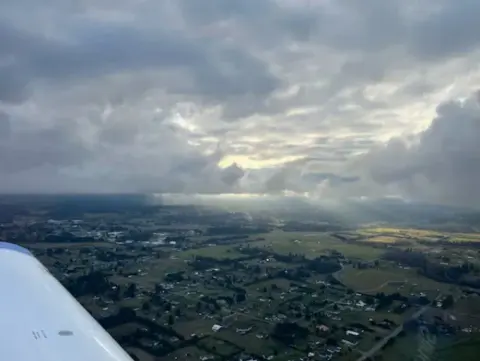 Rain showers over farmland seen past a wing