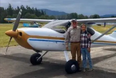 Man and a boy standing in front of a white and yellow taildragger