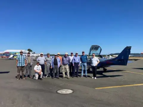 Fly-out group posing beside a low-wing airplane at Chehalis