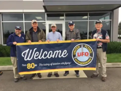 Group holding a Welcome banner in front of a hangar
