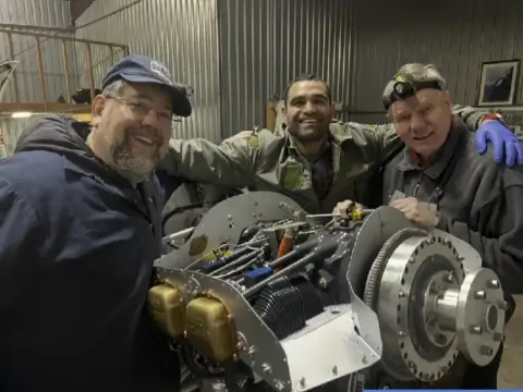 Three members smiling beside an airplane engine on a stand