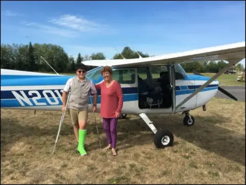 Emily and Aidan standing beside "Sally," a blue and white Cessna, on the grass
