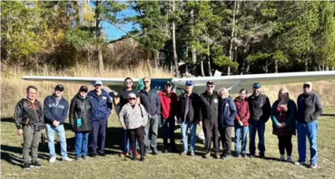 Roche Harbor fly-out group lined up on the grass in front of a high-wing airplane
