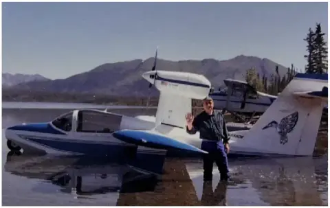An amphibian at the water’s edge with a pilot standing beside it and mountains behind