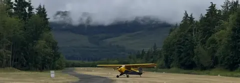 A yellow taildragger on a grass strip below cloud-covered hills