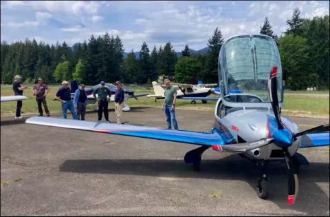 A blue and white low-wing airplane on the ramp at Packwood, with members talking in the background