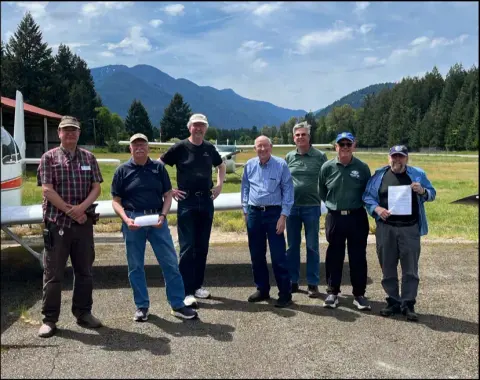 Smiling fly-out attendees lined up in front of a white low-wing airplane, with forested mountains behind