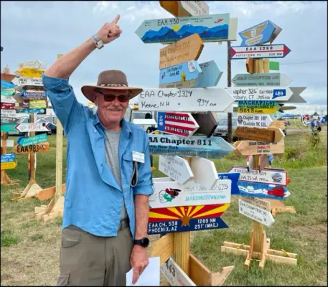 Man in a hat pointing at a signpost of destination arrows at a fly-in