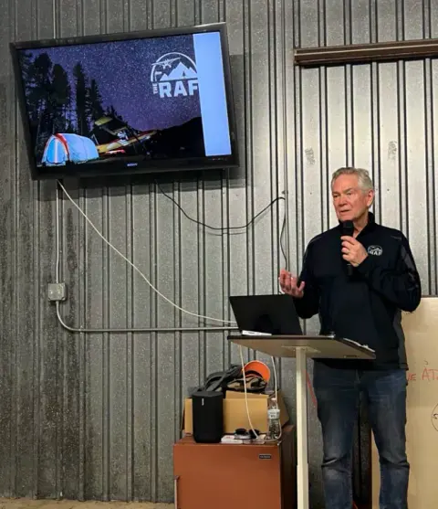 Speaker addressing members in a hangar beside a wall screen