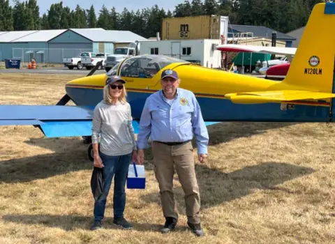 Skip and Ken Brown standing on the grass in front of a yellow and blue low-wing airplane
