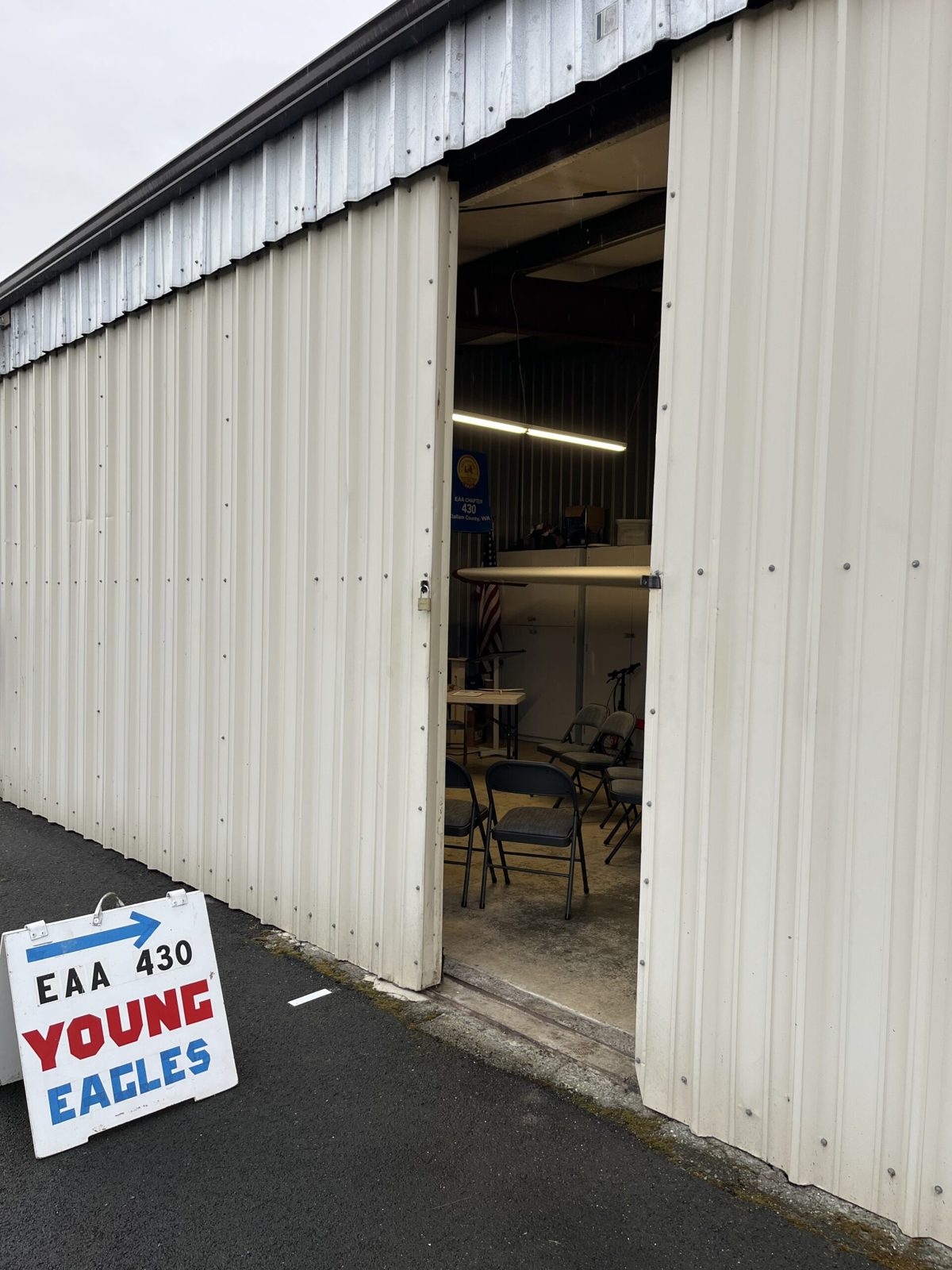 The open hangar door with a red, white, and blue sign on the pavement that reads EAA 430 Young Eagles, and folding chairs set up inside