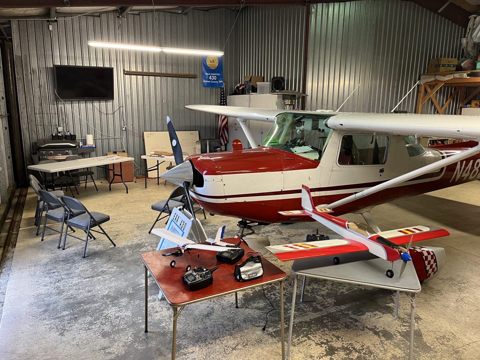 Inside the hangar, two radio-controlled model airplanes and their hand-held transmitters sit on small tables in front of a red and white Cessna