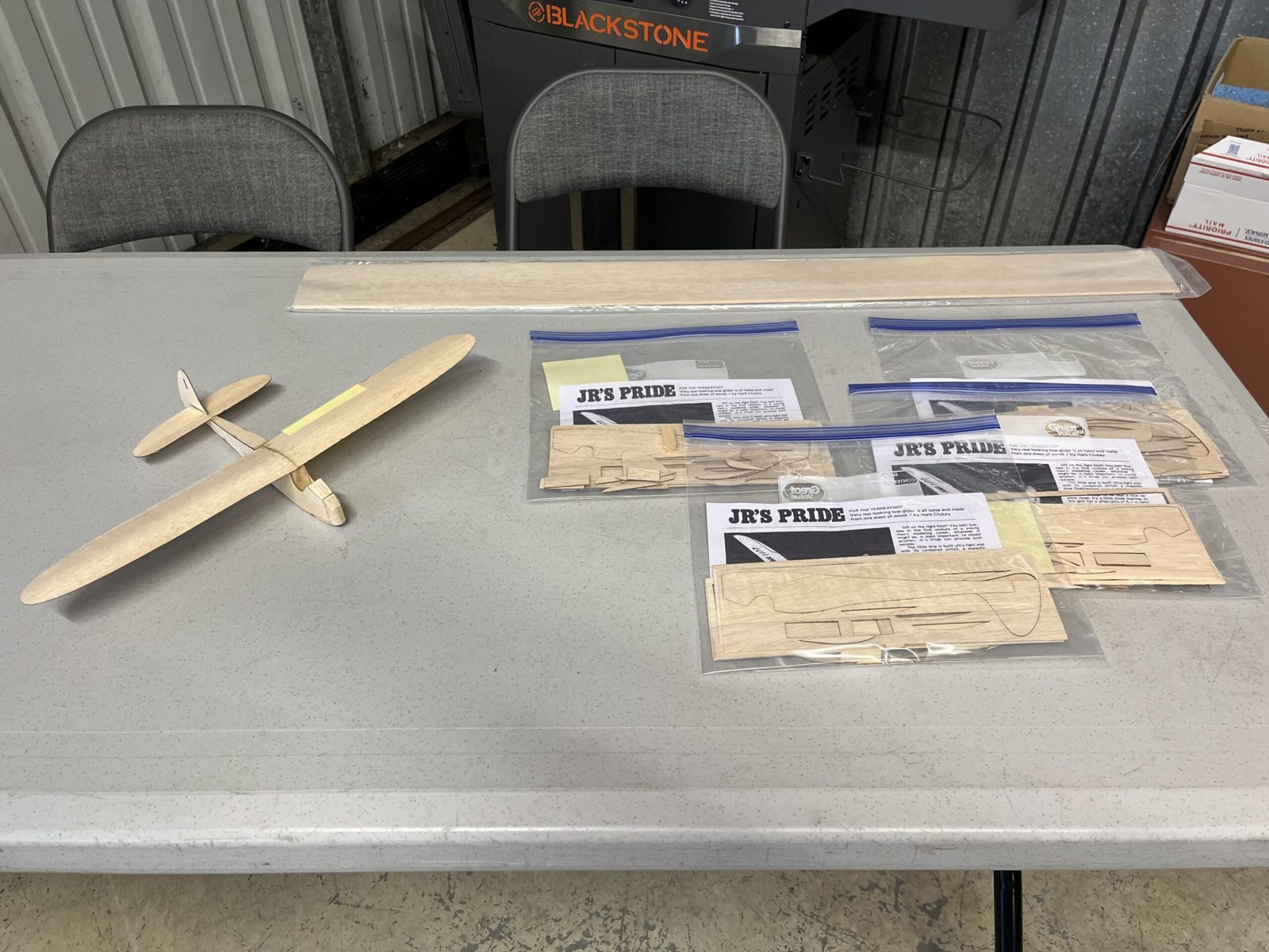 A finished small balsa wood glider rests on a table next to bags of glider kits labeled JR's Pride