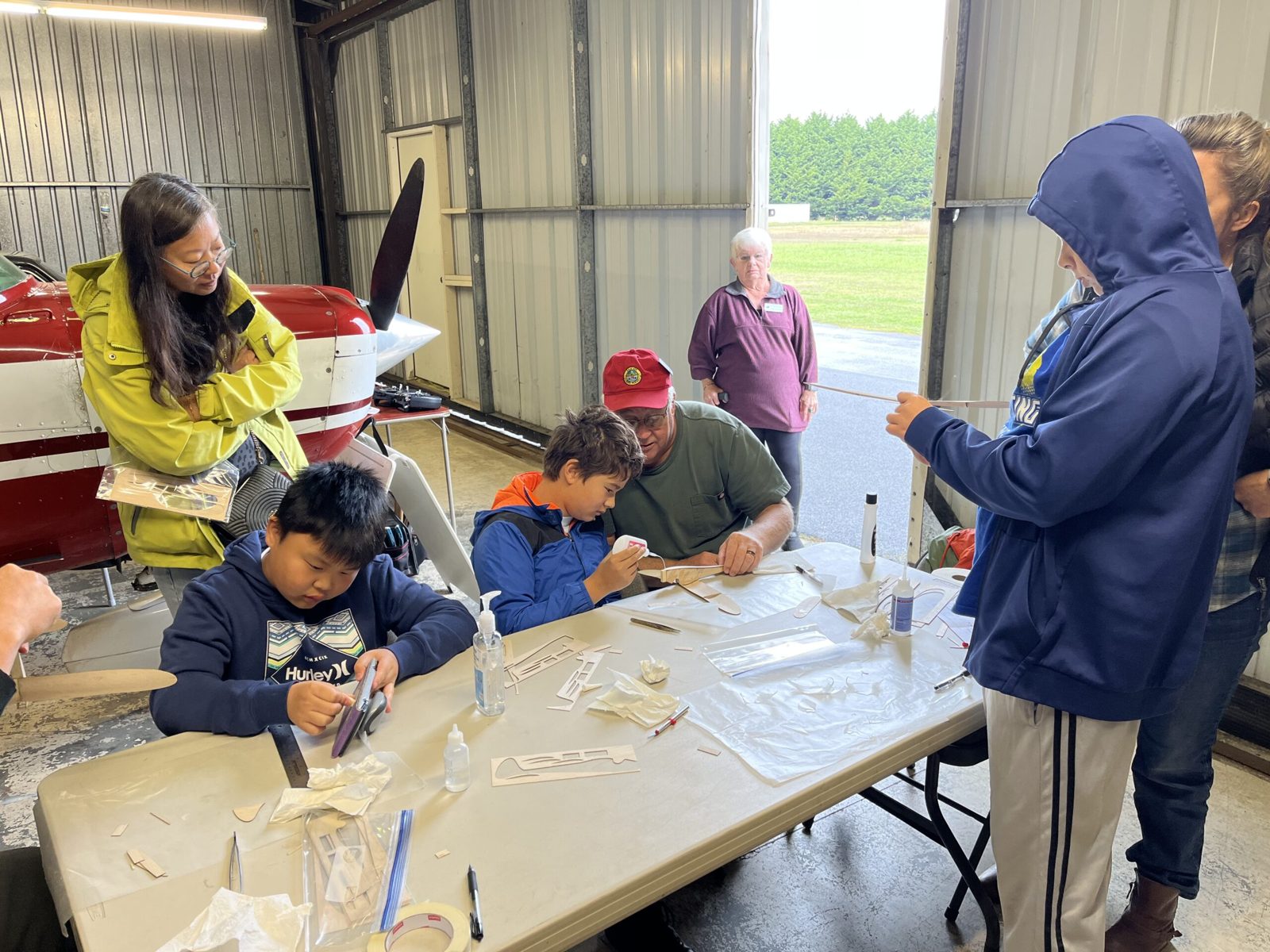 Kids and adult volunteers gather around a table in the hangar, gluing and assembling balsa glider parts