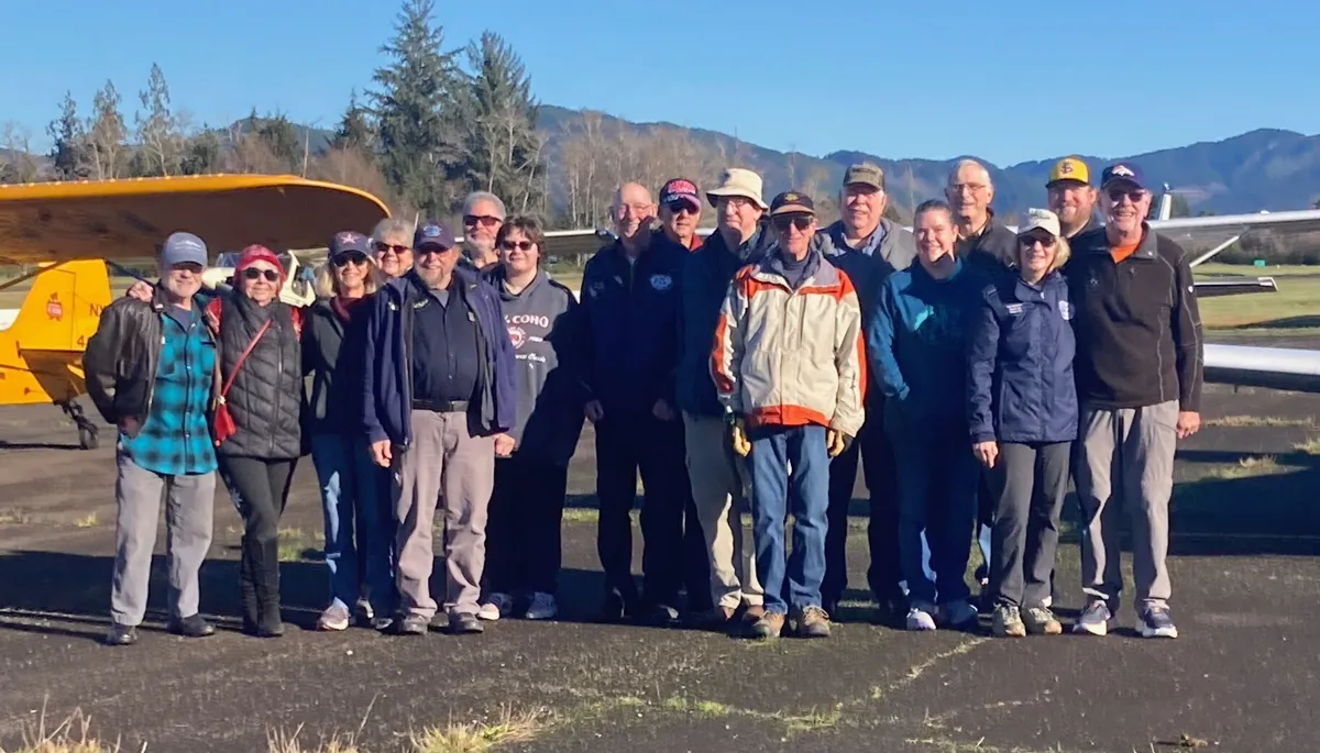 About twenty EAA Chapter 430 members posing for a group photo on the ramp during the January 2026 fly-out to Forks, with a yellow high-wing airplane, tall evergreens, and foothills behind them.