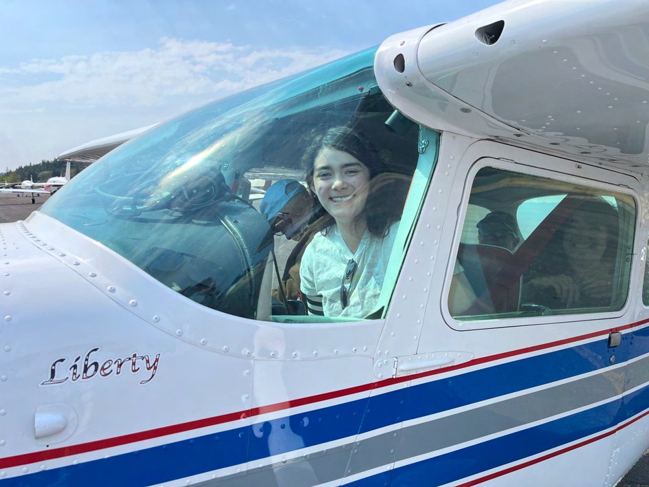 A smiling young pilot in the left seat of a white high-wing Cessna with passengers aboard