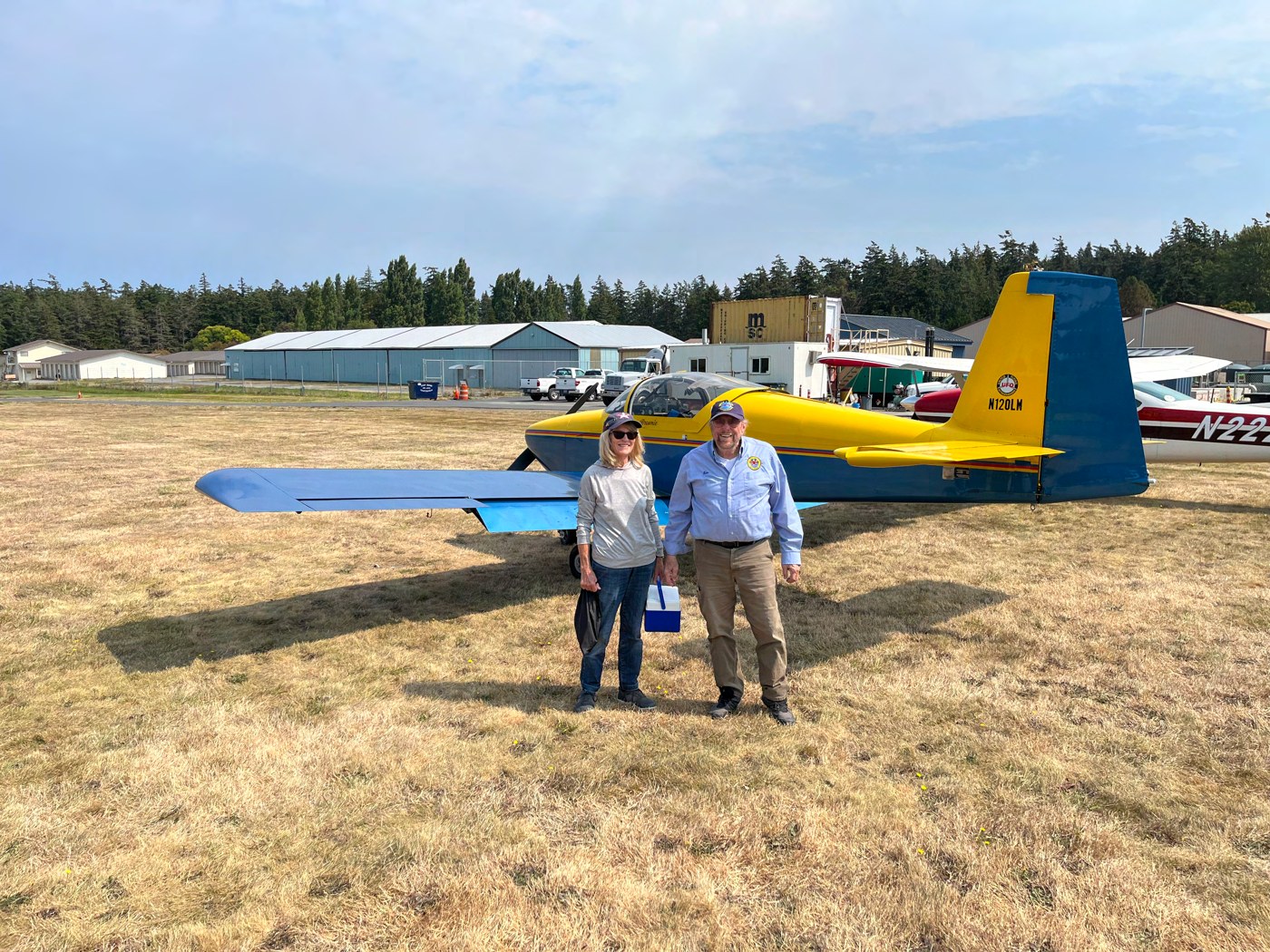 Two fly-out attendees standing on the grass in front of a yellow and blue low-wing homebuilt airplane